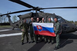 Service members wearing flight suits hold a Slovak flag while standing next to a military helicopter.