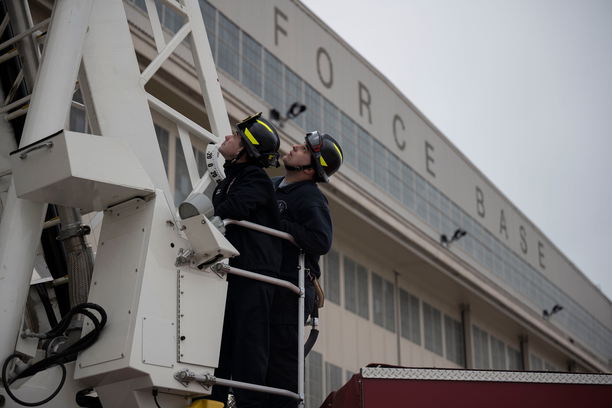 Two firemen in black uniforms and helmets look up at the extended firetruck ladder.