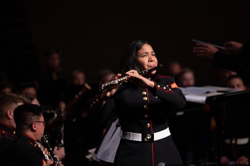 A Marine plays the flute on a dark stage with an orchestra behind her.
