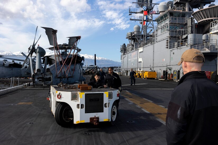 Sailors stand on the deck of a ship with a cart and a helicopter in the background.