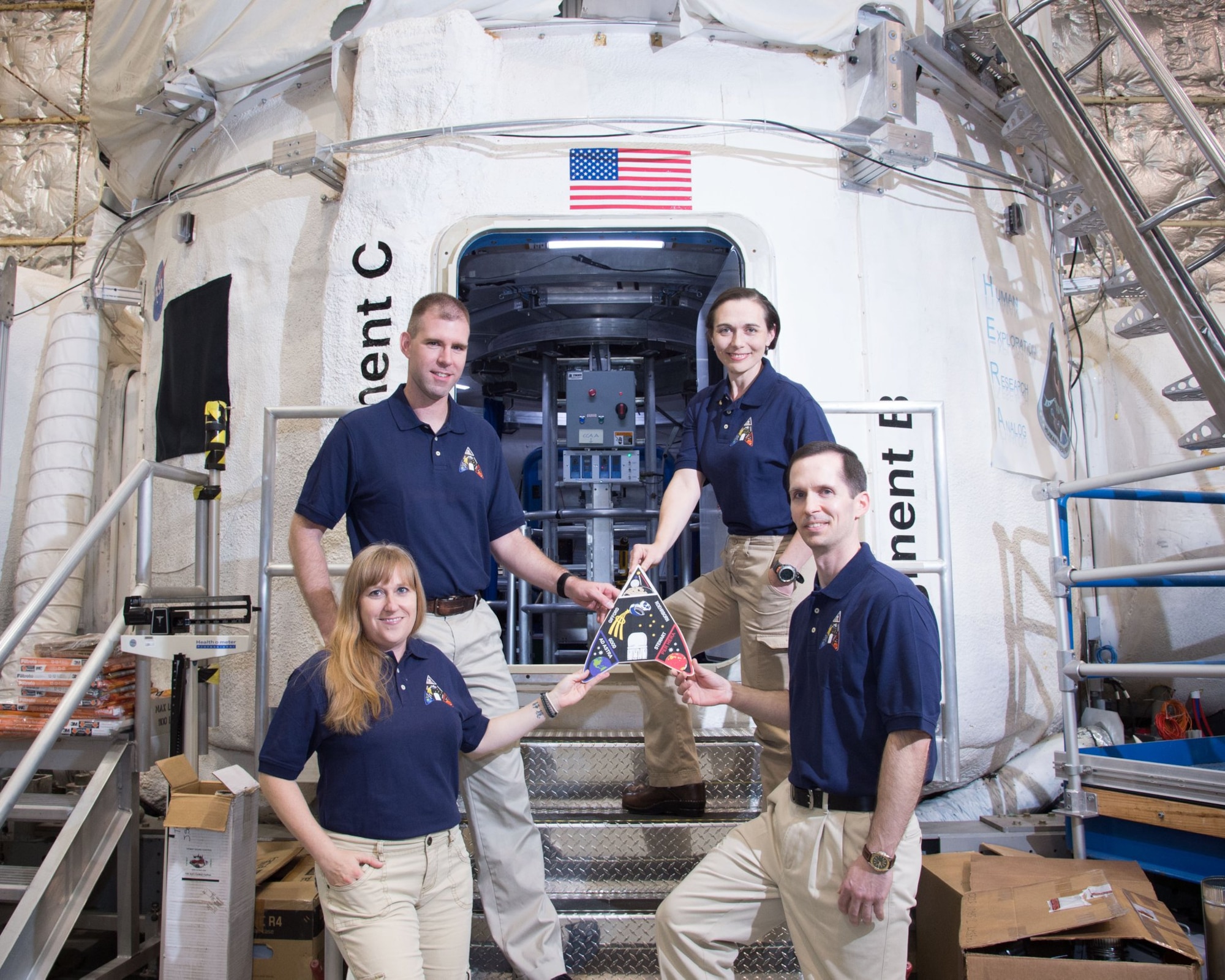 Four people stand in blue polo shirts and khaki pants. Two are standing in each row, all in front of a space craft vessel.