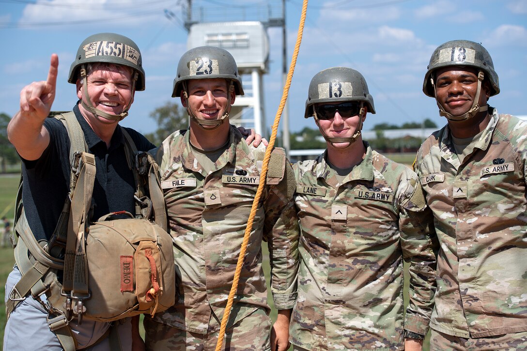 Three service members wearing hard helmets pose in line for a photo with a civilian holding up a forefinger.