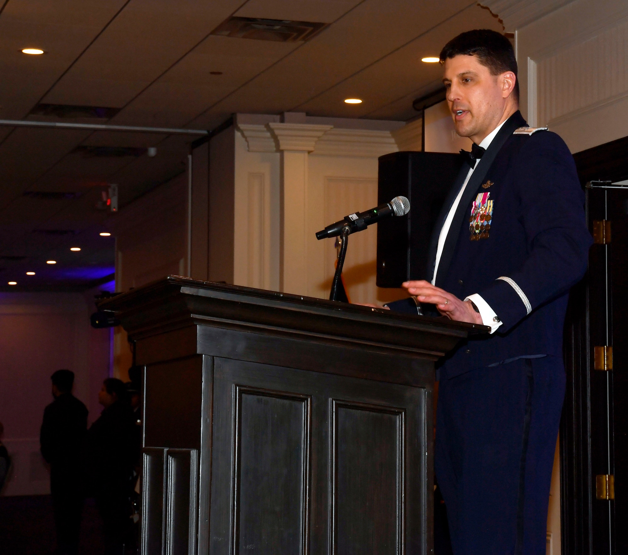 Male servicemember in formal dress stands behind podium on a stage.