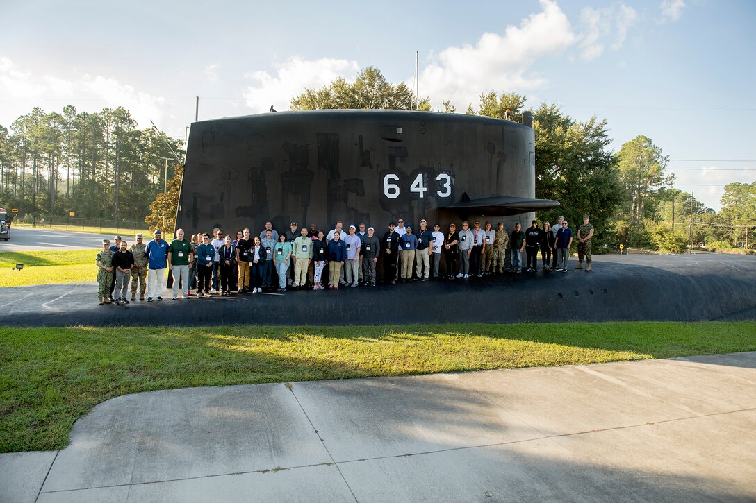 A large group of service members and civilians poses in front of an outdoor submarine display with trees in the distant background.
