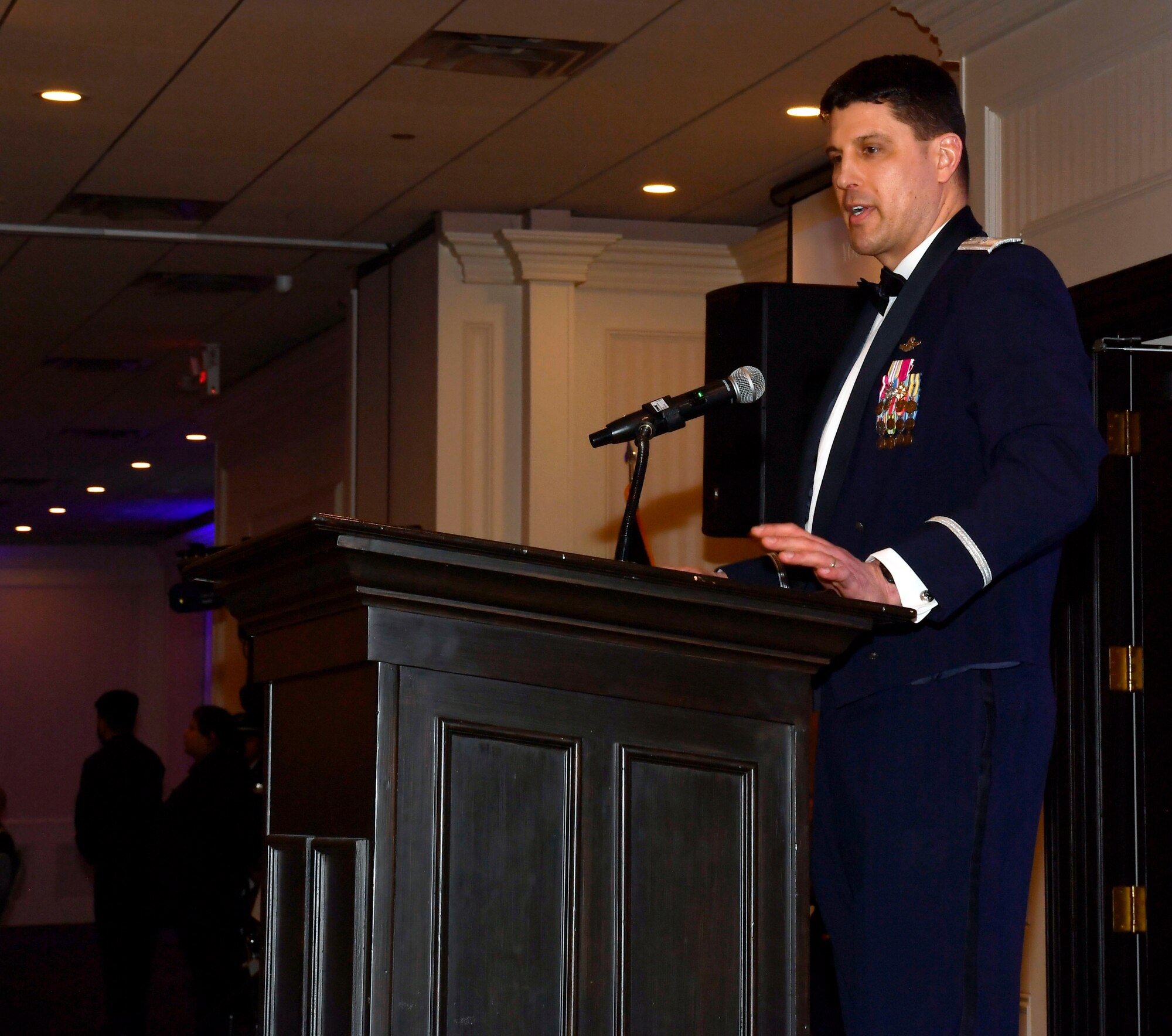 Male servicemember in formal dress stands behind podium on a stage.