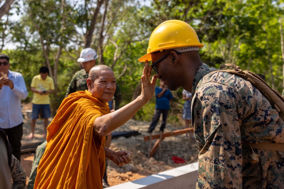 A monk places his hand in front of the face of a Marine wearing a hard hat.