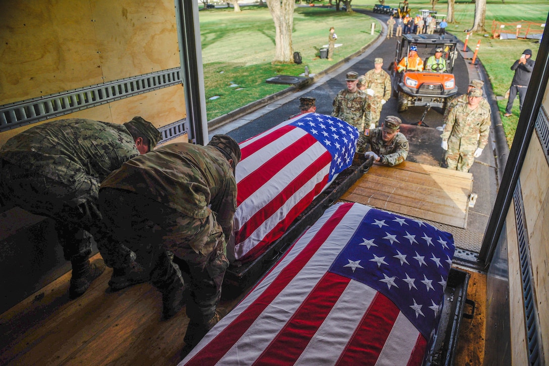 Service members place two caskets covered by American flags into a vehicle while other service members stand at attention on a road. Other vehicles are parked in the background.