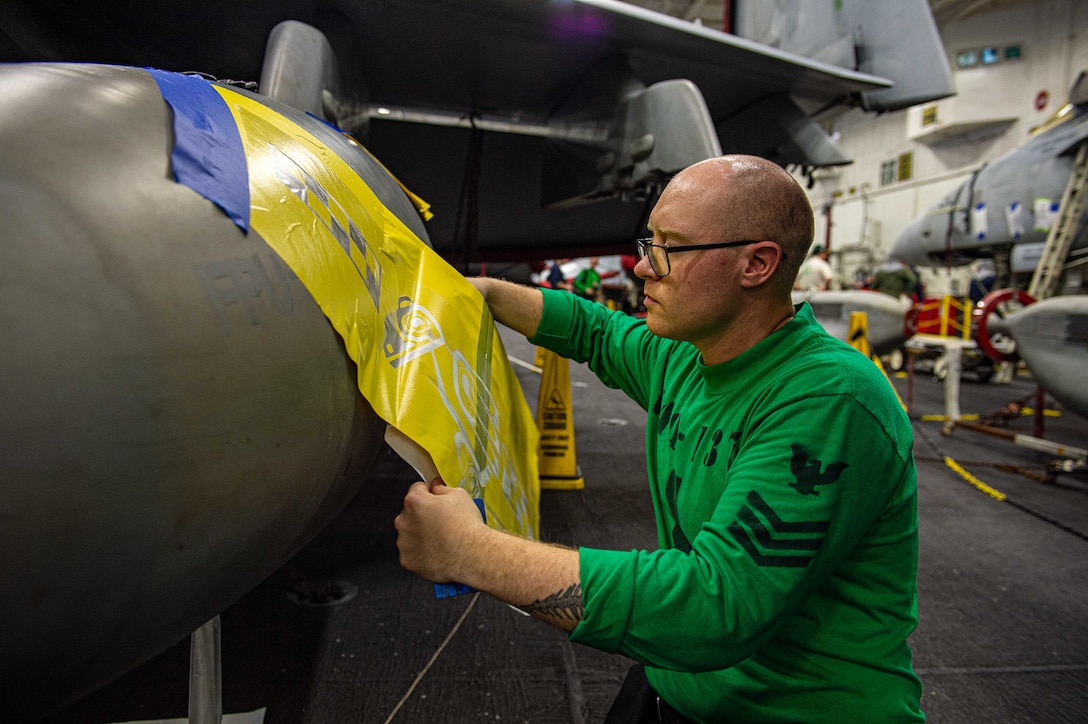 A sailor kneels to apply a decal to a military aircraft inside a ship’s hangar bay.
