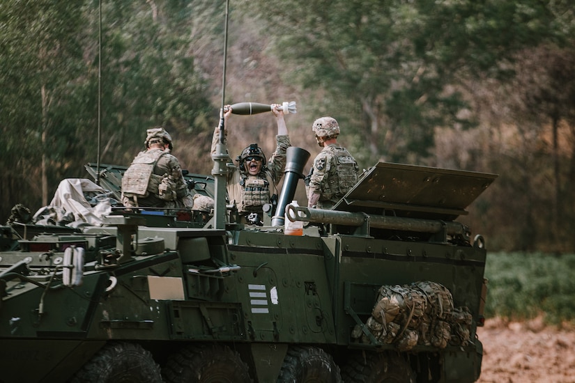 A soldier holds up a mortar round during training as others sit inside a military vehicle.