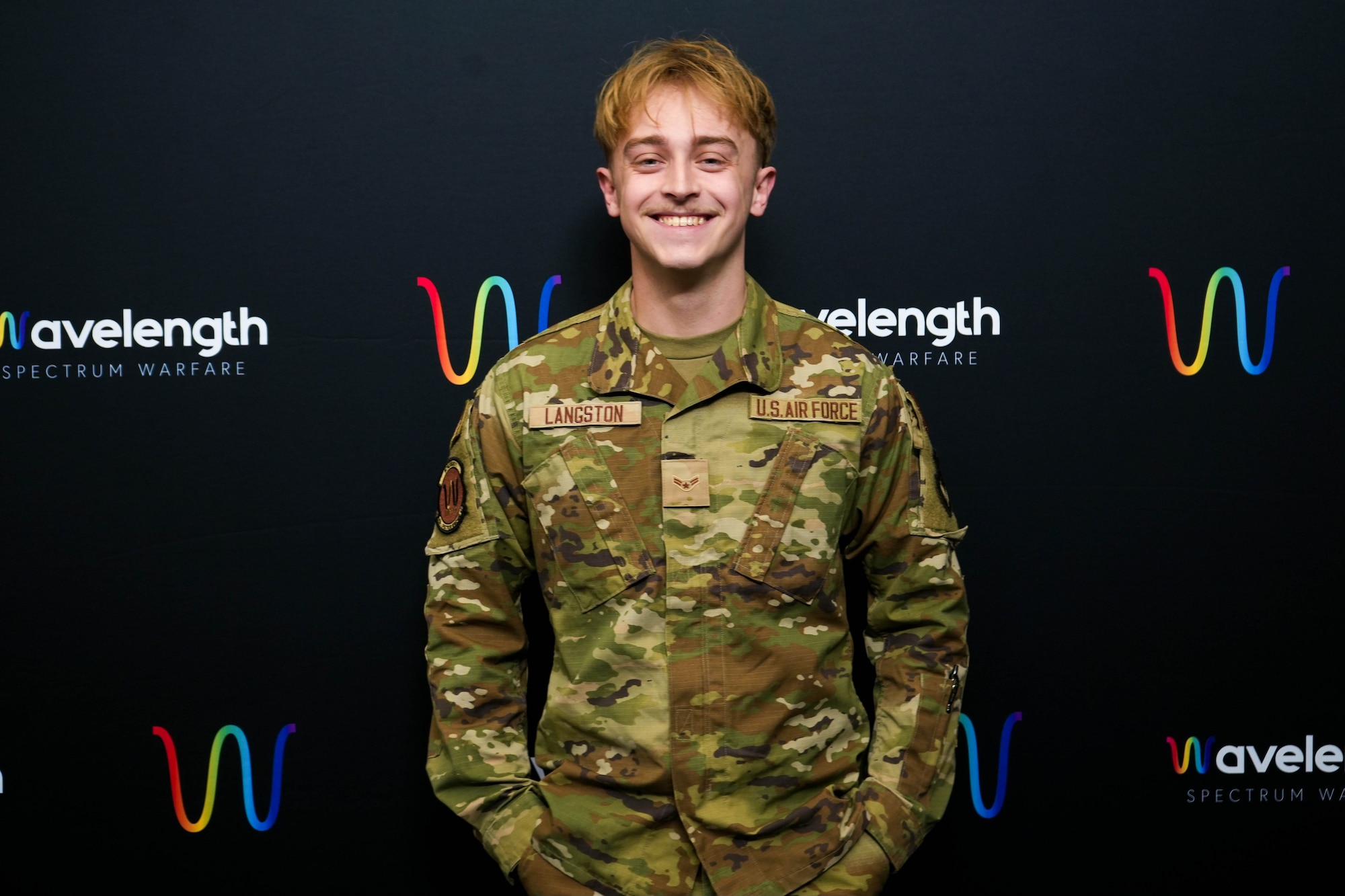 Airman First Class Kaden Langston, 850th Spectrum Warfare Group, Detachment 1, software designer, poses for a photo in front of his detachment logo in San Antonio, TX, March 4, 2024. The mission of the detachment is to experiment, measure, and learn with fully-kitted teams to deliver Electromagnetic Spectrum Warfare mission focused software. (Courtesy Photo)