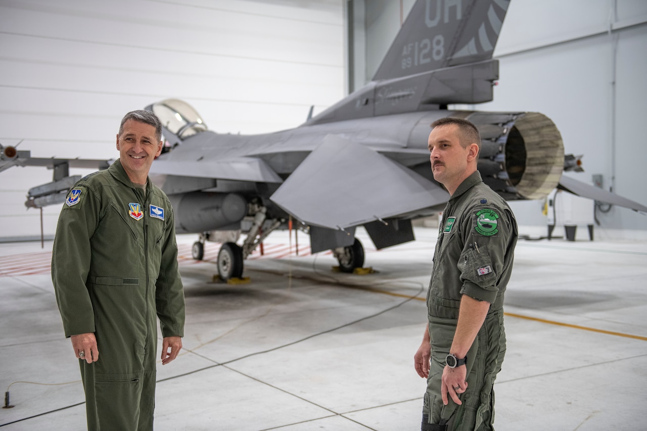 Two men chat in front of and F-16 fighter jet.