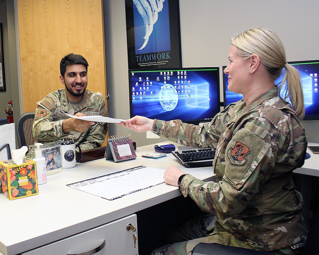 Two Airmen at a desk, handing a piece of paper from one to another.