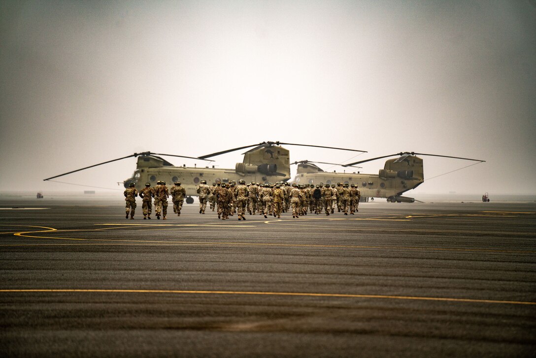 A group of uniformed service members walk toward a military aircraft on a cloudy day.