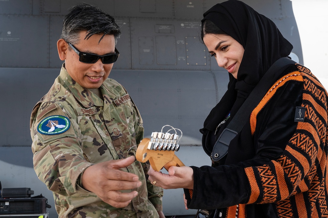 A civilian holds a guitar as an airman gestures toward the top of the instrument.