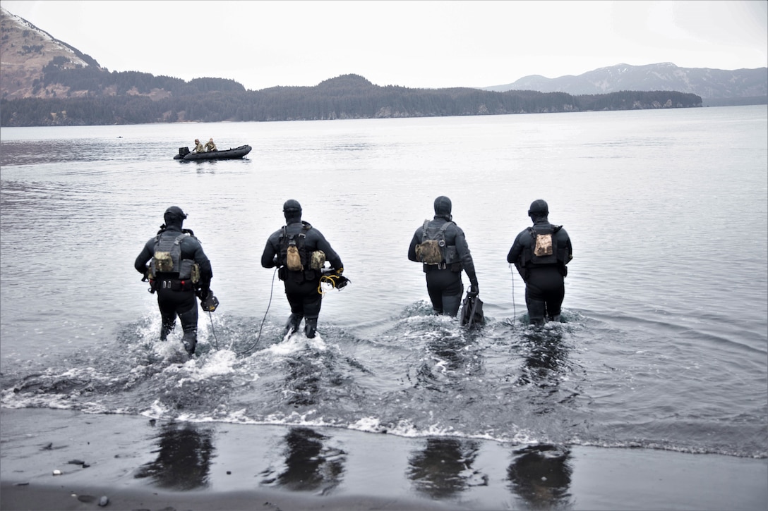 Four service members in diving gear walk into water as two other people are in a raft further out in the water. Mountains can be seen in the distance.