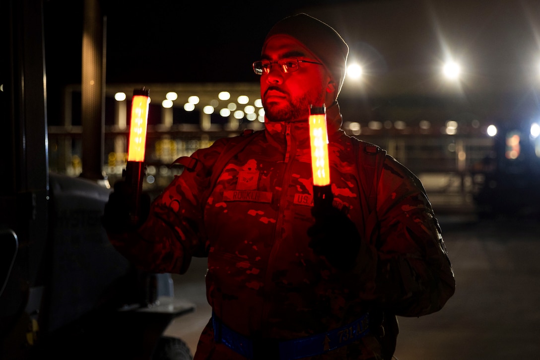 An airman holds two batons illuminated by orange light while standing next to a truck as spotlights shine in the background.