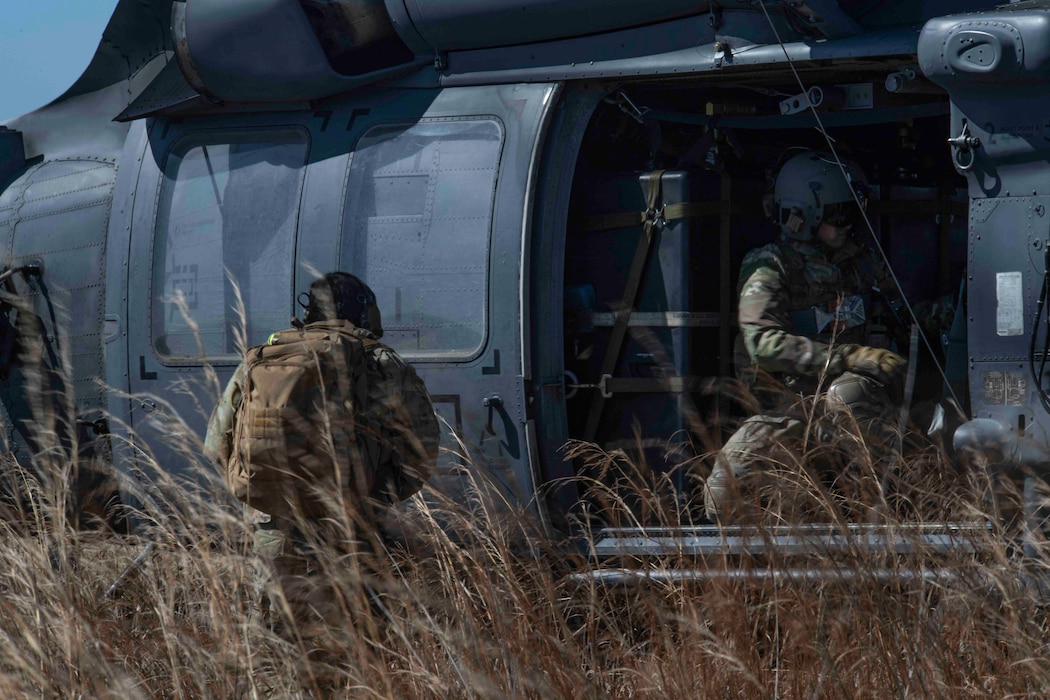 A man in camouflage fatigues with a tan backpack approaches a helicopter's open side door, where another man in a flight suit and helmet waits to help him board.