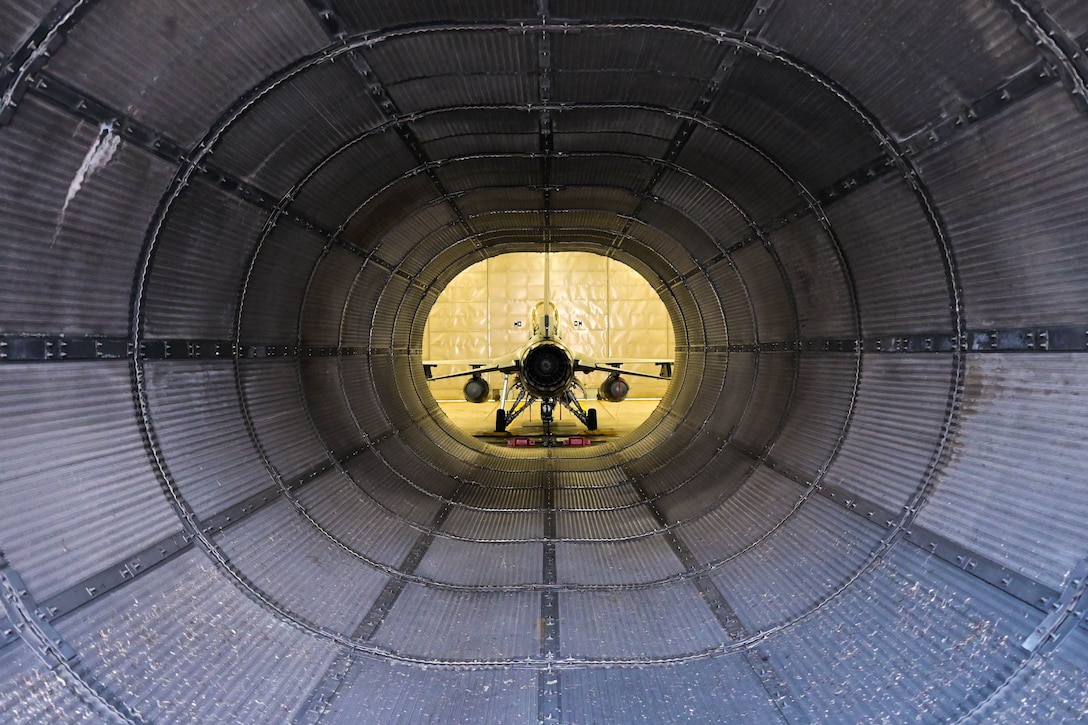 The rear of a fighter jet is seen through a large metal tube.