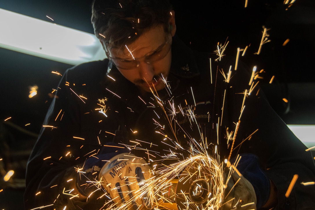 Sparks fly as a sailor wearing gloves and protective eyeglasses uses a circular saw to cut a piece of metal.