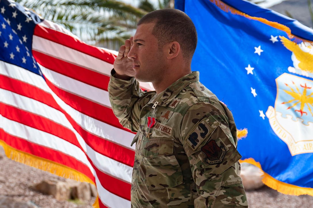 Close-up of an airman saluting with flags in the background. The airman is facing the left.