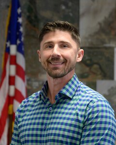 A man smiles while standing in front of an American flag and a grid map