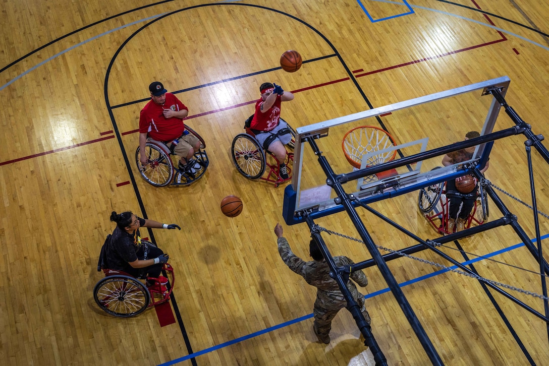 A service member standing under a basketball net passes a basketball to one of four veterans sitting in a wheelchair as another takes a shot.