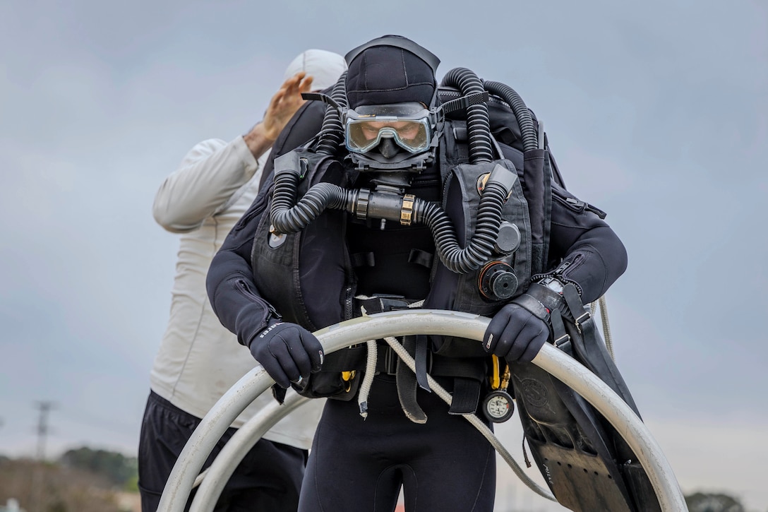 A sailor wearing a wetsuit and underwater breathing apparatus holds a rail as someone adjusts their equipment from behind.