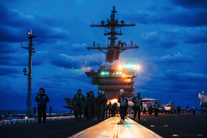 Sailors walk in formation aboard a ship under a dark, cloudy sky as spotlights illuminate the background with green and orange lights.