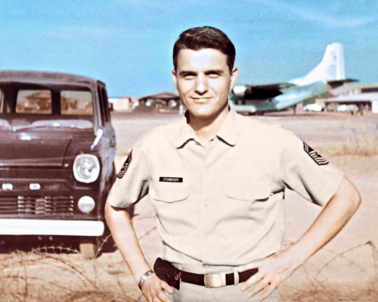 A man poses for a photo with his hands on his hips. An airplane is in the desert background.