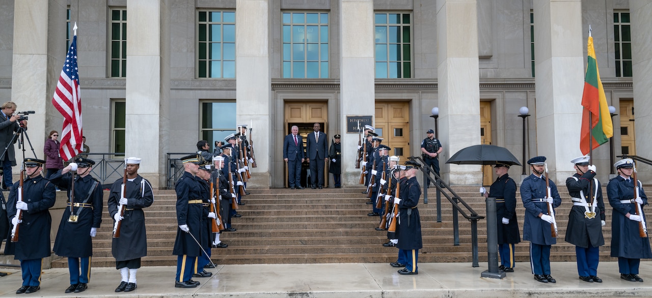 Secretary of Defense Lloyd J. Austin III stands with another defense minister at the top of steps with honor guards holding rifles and flags.