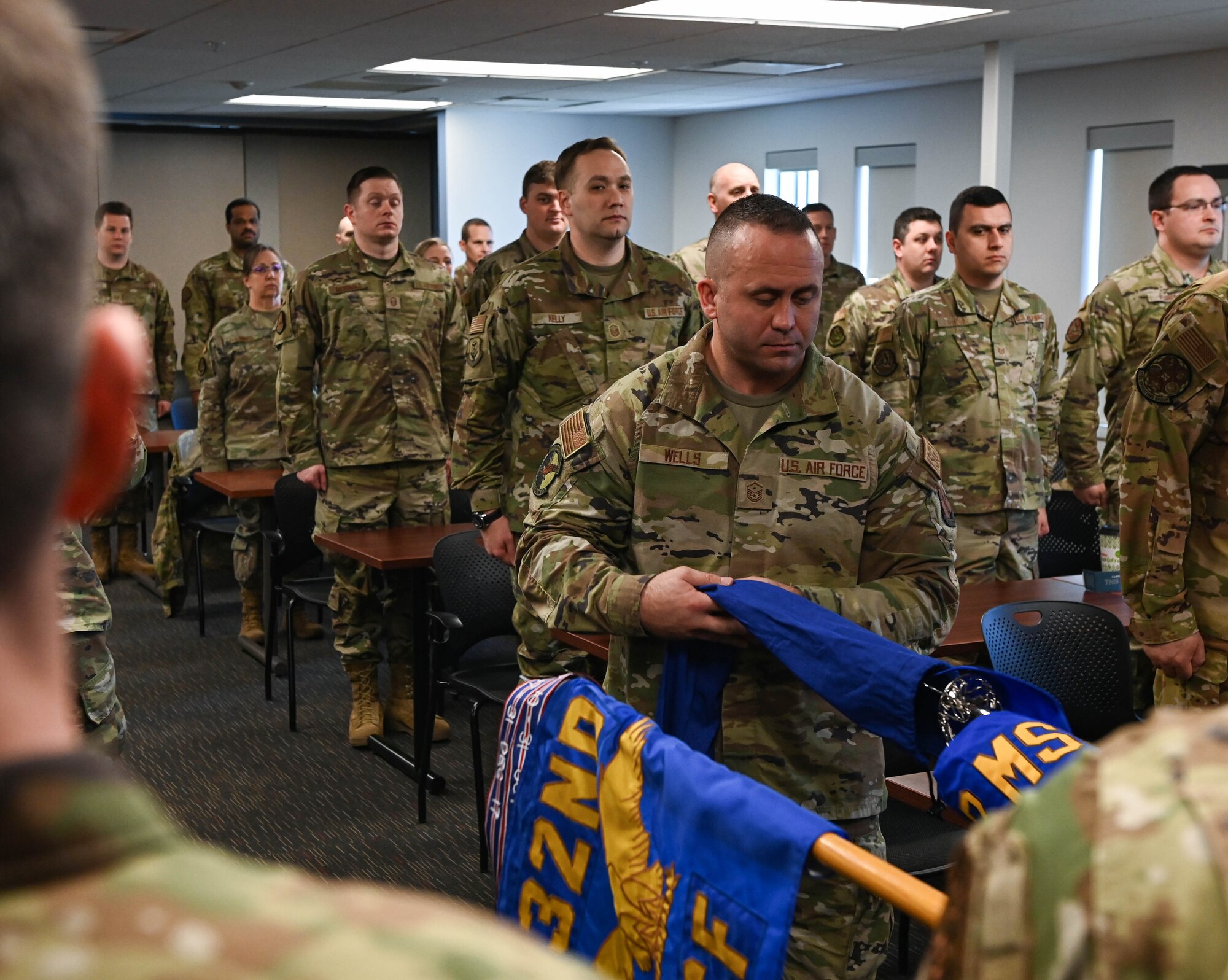 Man in camo uniform removes covering from blue flag with a crowd of Airmen watch