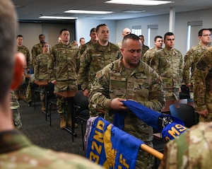 Man in camo uniform removes covering from blue flag with a crowd of Airmen watch