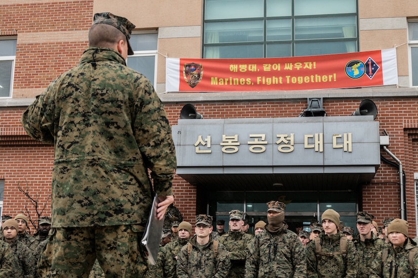 A Marine officer speaks to a large group of Marines in front of a building.