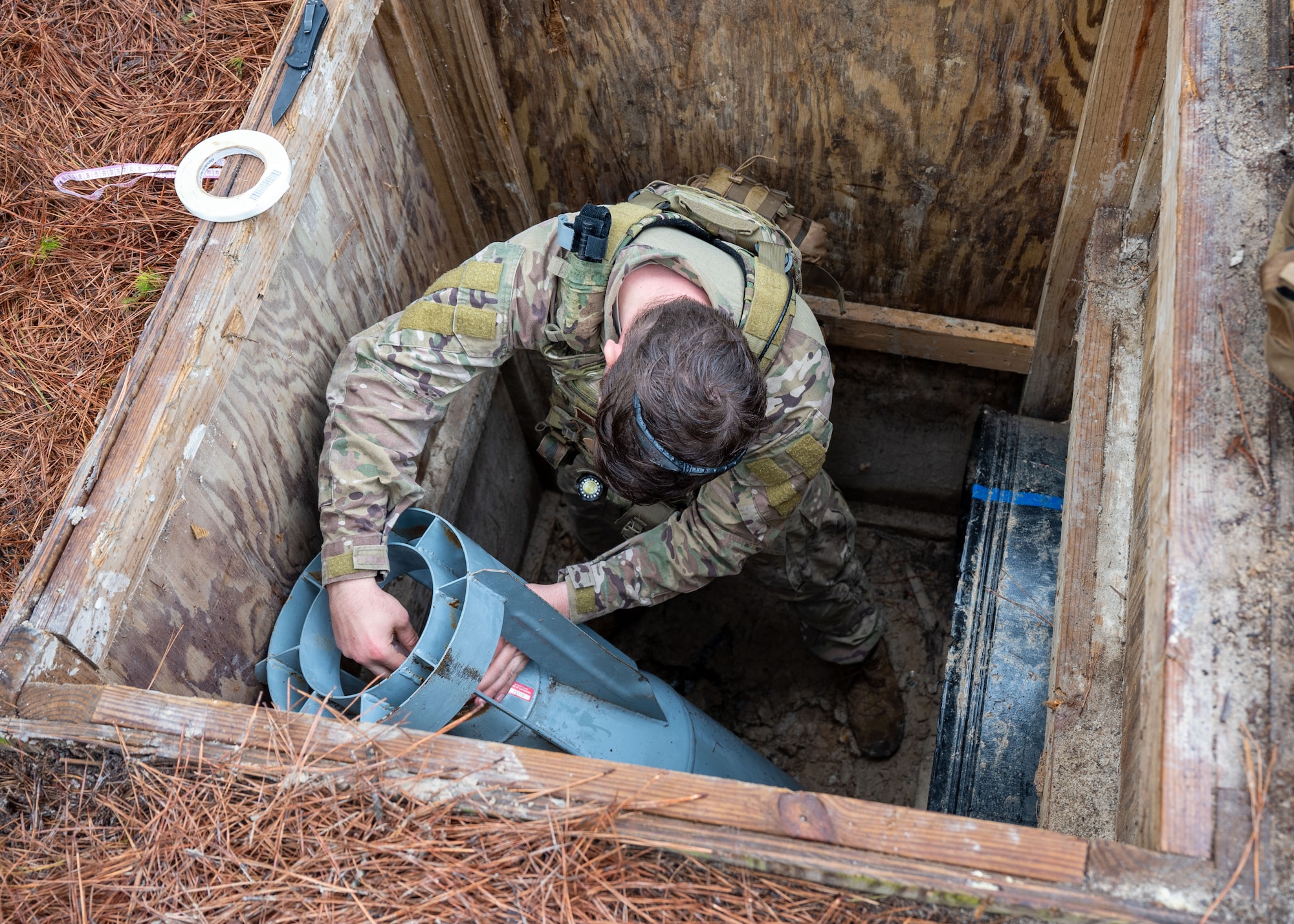 A man in a hole examines a training ordnance within the hole.