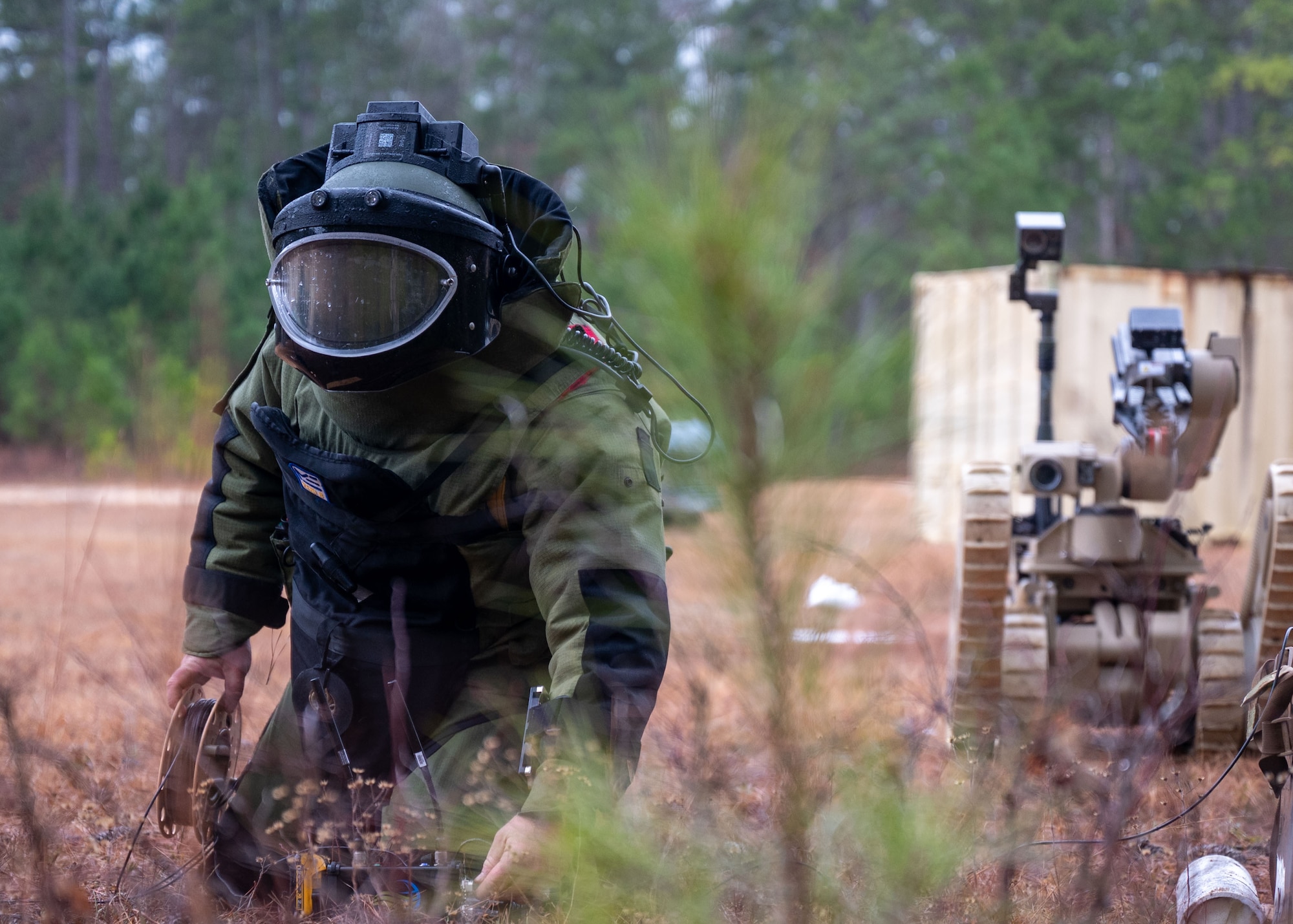 A man in an EOD protective bomb suit kneels in the grass.
