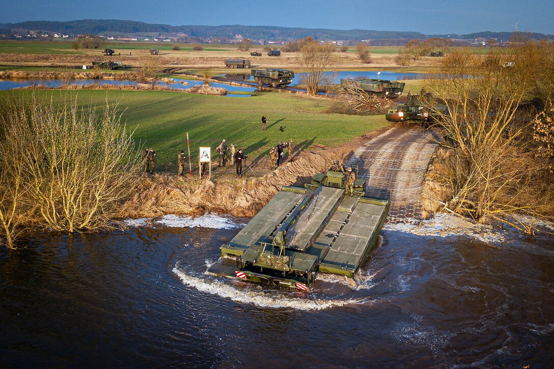 A flat-topped amphibious vehicle makes a splash as it enters a body of water from a dirt road where other vehicles are moving.
