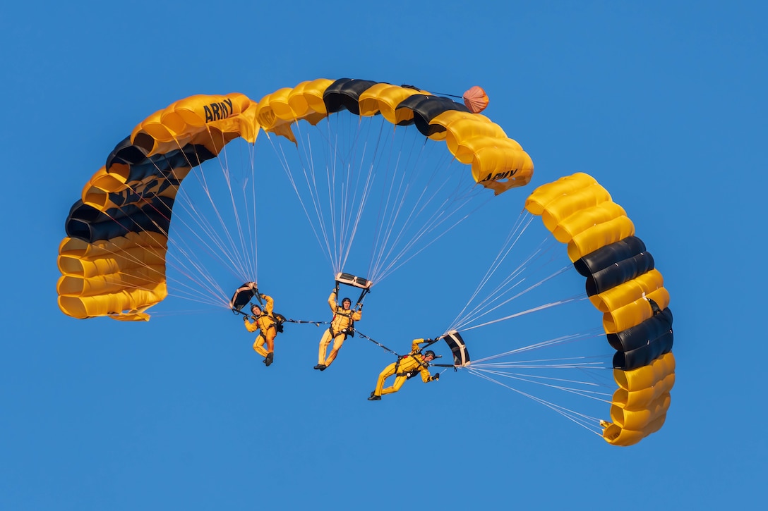 Three soldiers tied together parachute to the ground against a blue sky.
