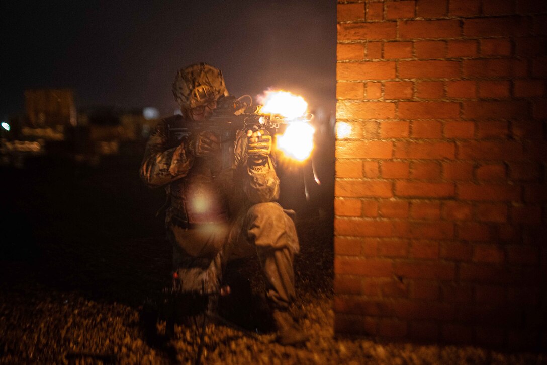 An airman kneels and fires his weapon next to brick wall at night.