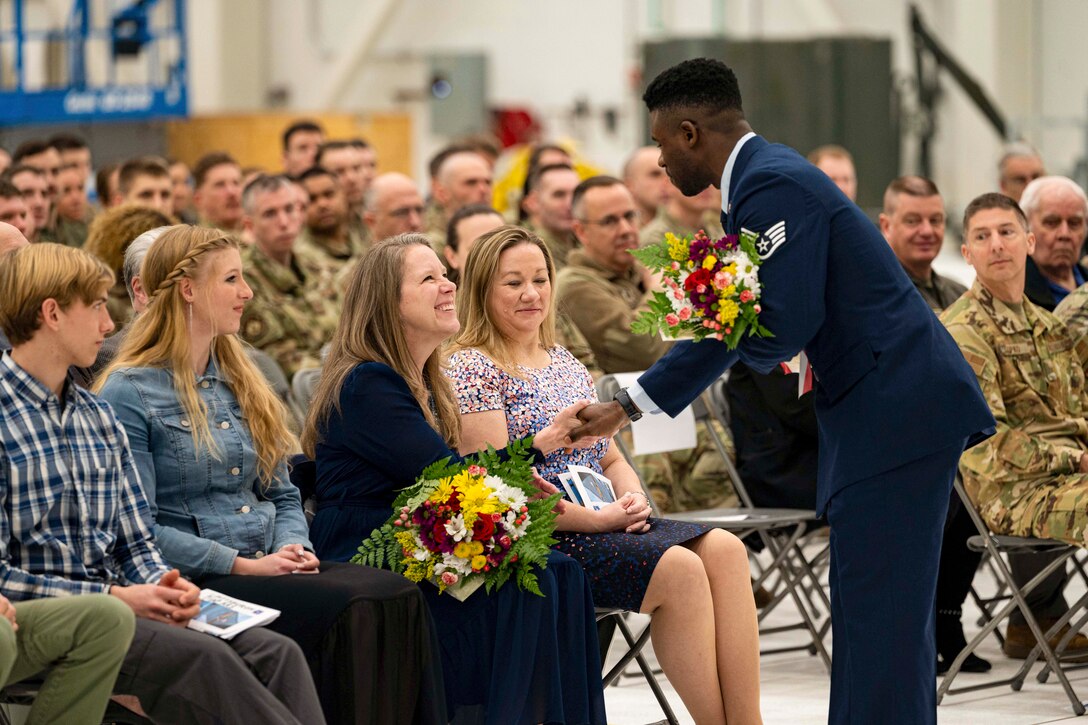 An airman and a civilian each holding flowers shake hands as others sitting in an audience watch.