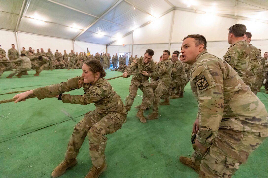 ROTC cadets pull a rope during a competition as fellow cadets cheer them on from the sideline in a tent.