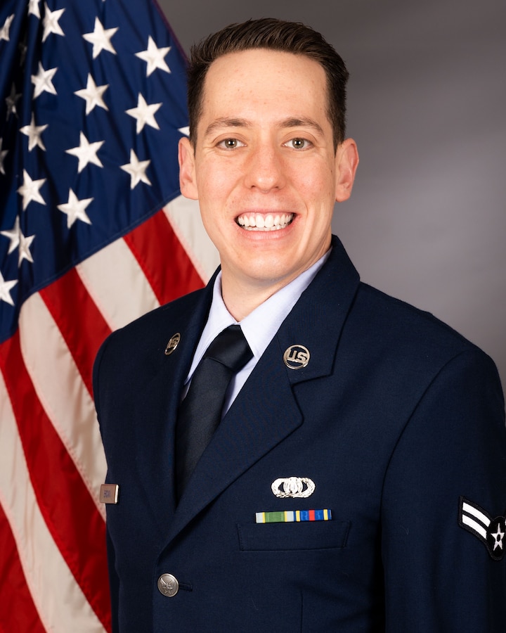 Official photo of A1C Steven brown in front of the American flag. He is wearing his blue service dress uniform.