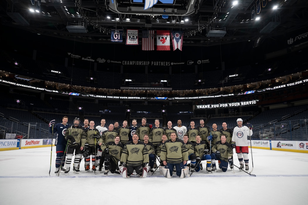 Hockey players smile for a photo wearing military green and black Columbus Blue Jacket jerseys.