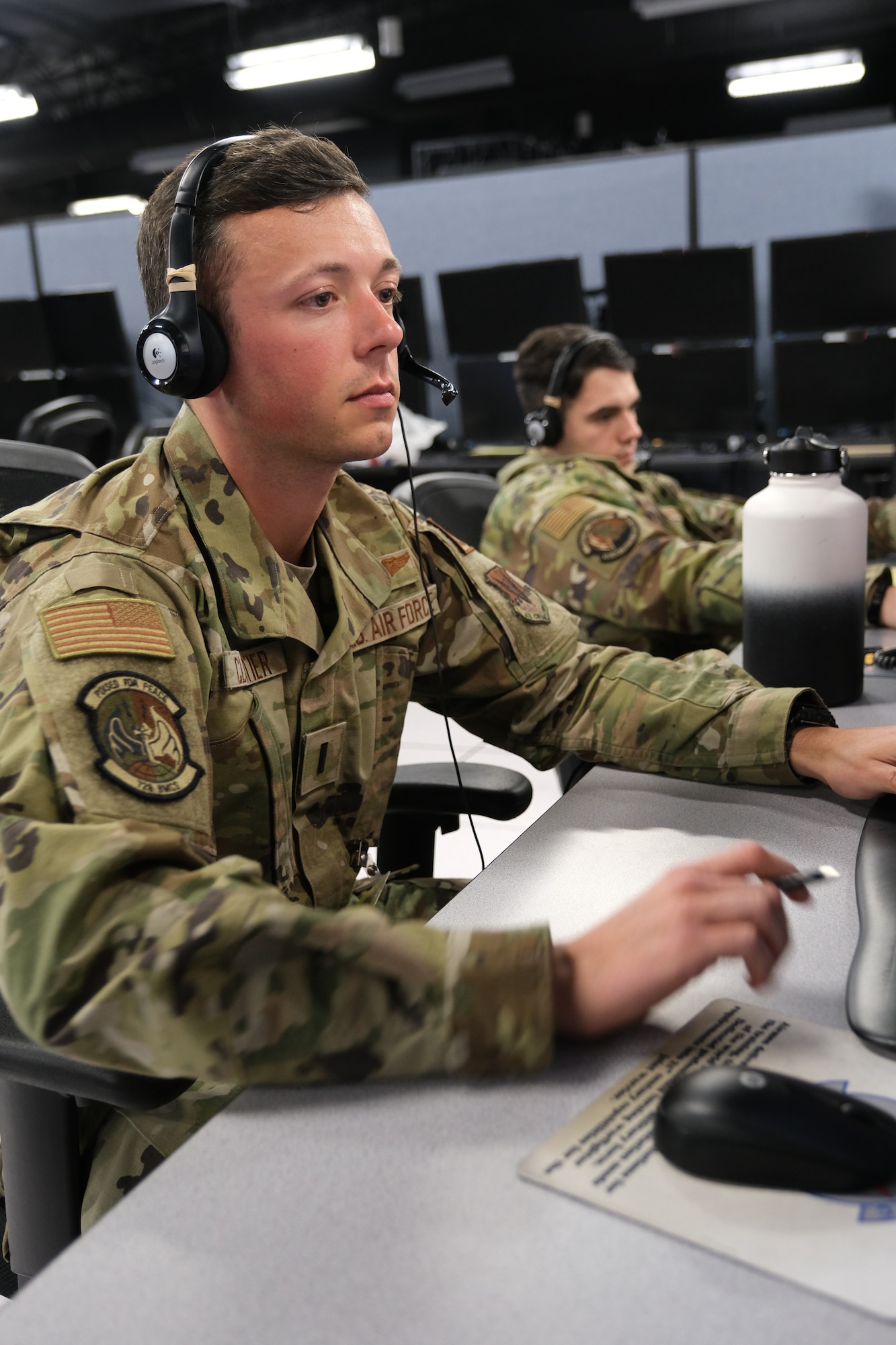 two uniformed U.S. Air Force Airmen wearing headsets, work at computers, in the background are multiple computer screens