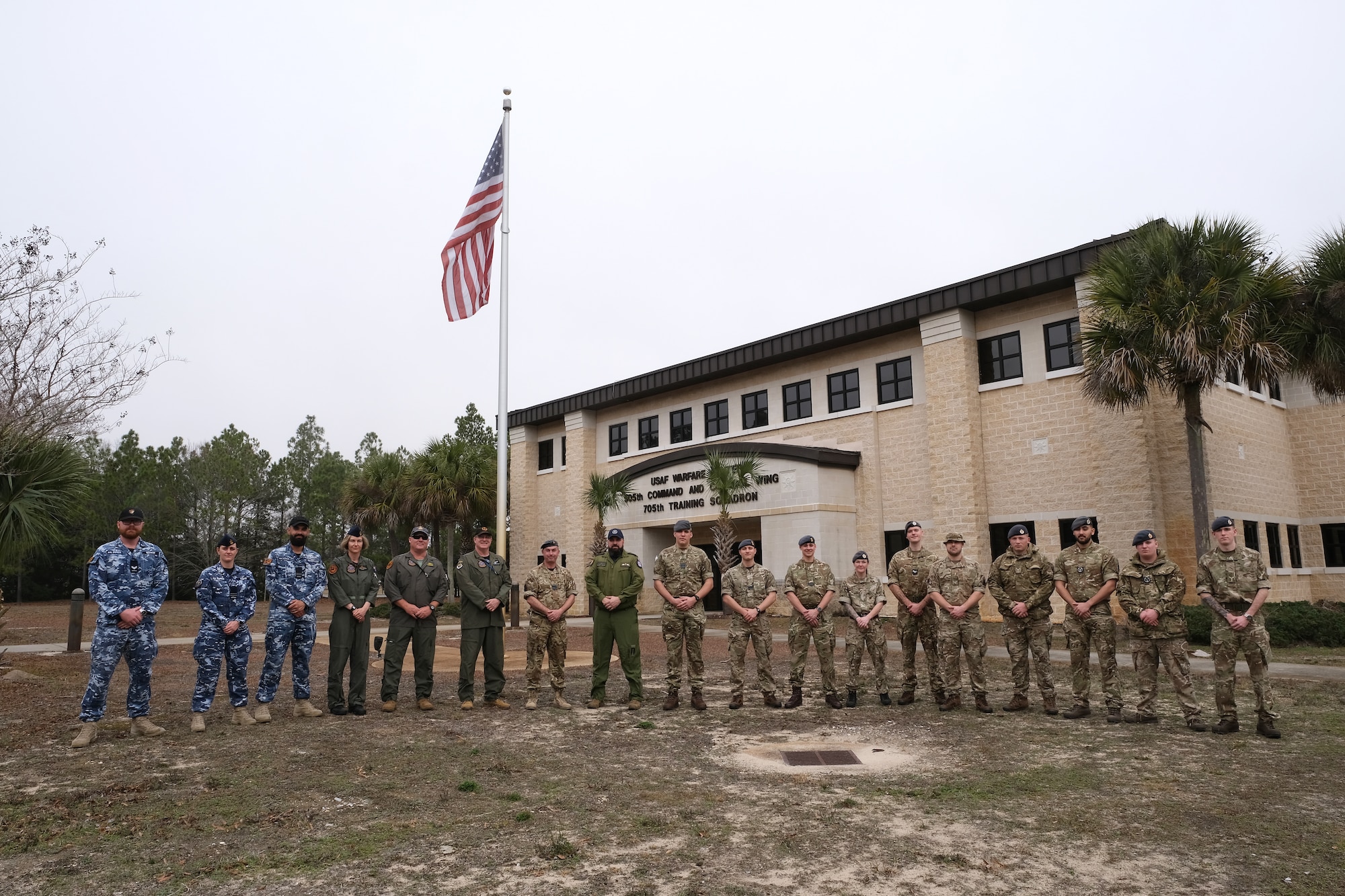 United Kingdom, Royal Air Force, Royal Australian Air Force, and Royal Canadian Air Force members stand outside a building with a U.S flag flying in the background.