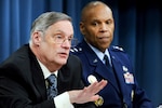 A person in a business suit speaks as a uniformed service member watches.