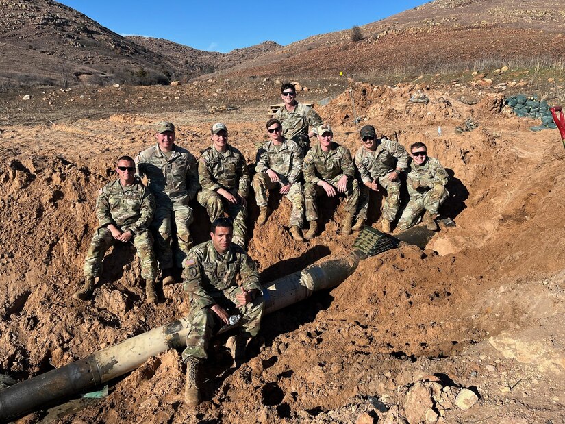 A group of soldiers stand and sit in a dirt pit and pose for a photo with a jammed round while one sits on top of it.