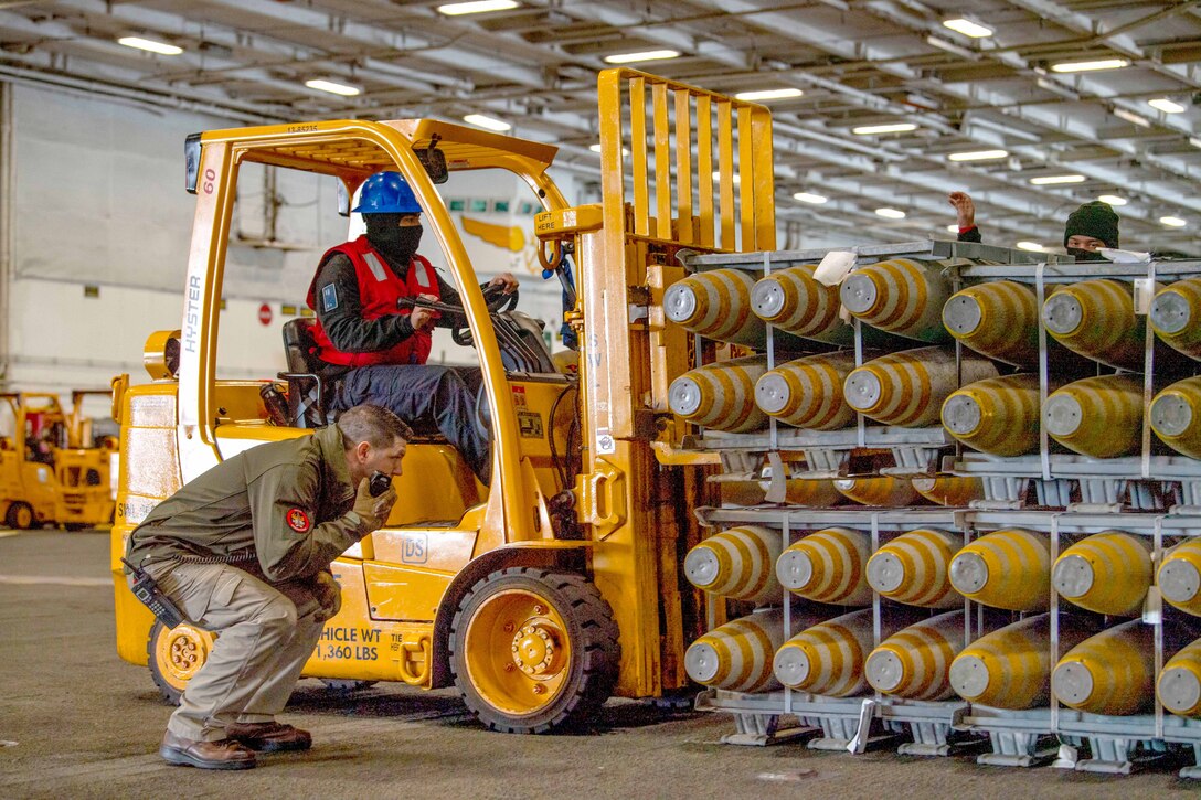 A sailor uses a forklift to move stacks of ammunition while another sailor crouches and holds a hand-held device.