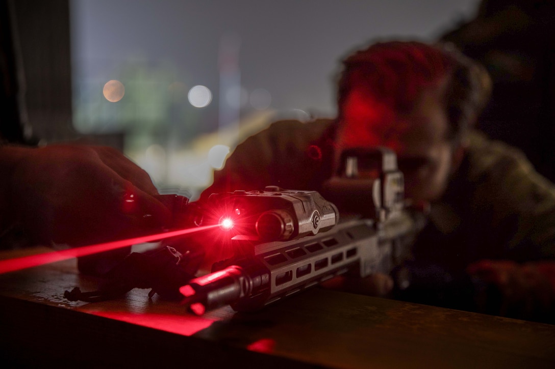 A soldier aims a weapon, which shoots out a red laser beam in a darkened area.