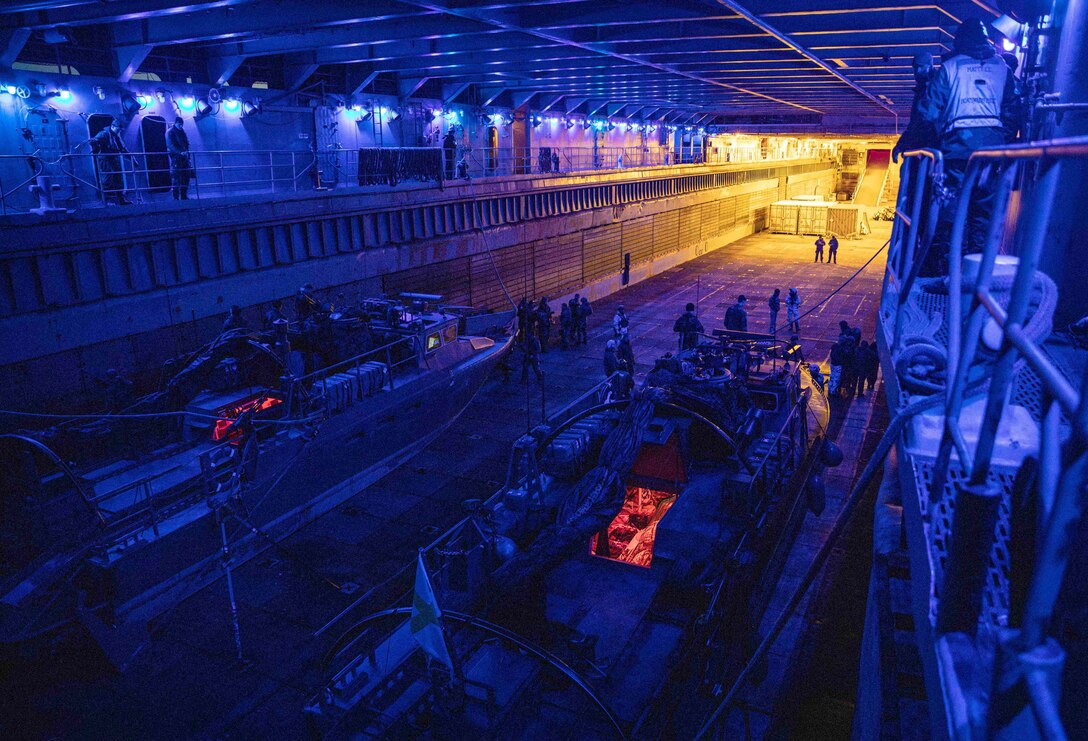 Service members stand around two small boats in a darkened well deck of a ship under purple lights.