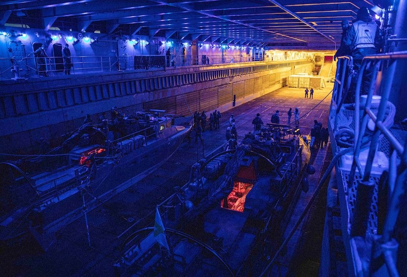 Service members stand around two small boats in a darkened well deck of a ship under purple lights.